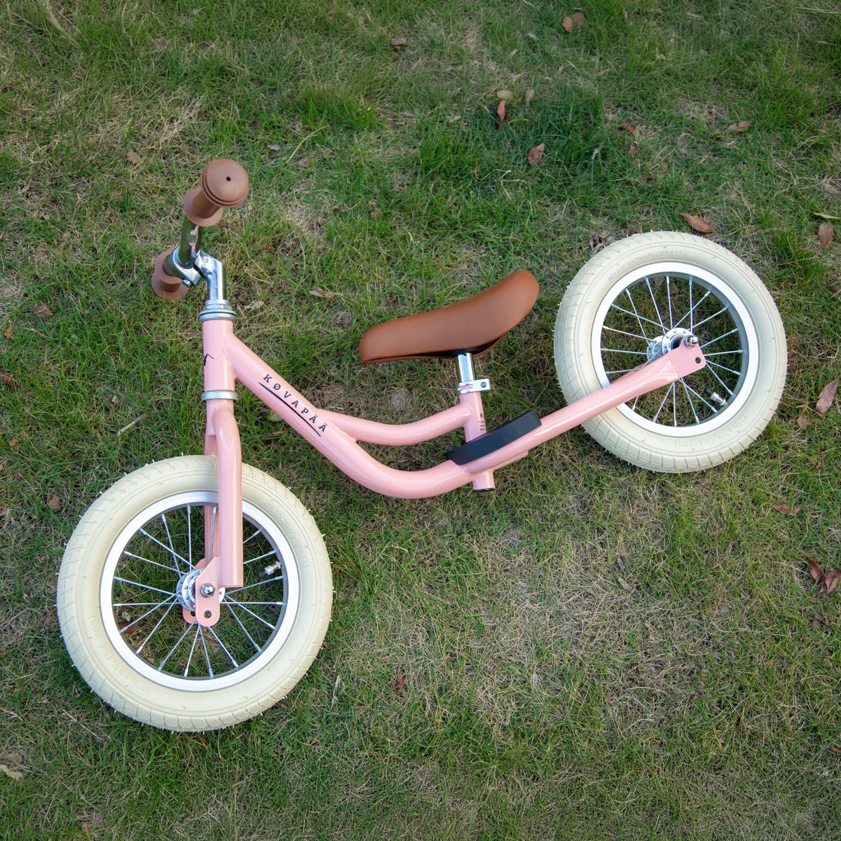 Kovapää 2 in 1 Loopfiets Roze met bruine zitting en witte banden op gras.